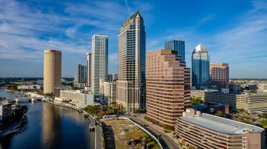 Tampa, Florida 'nın hava manzarası modern yüksek binalarla dolu canlı bir ufuk çizgisi, şehir merkezinden geçen ikonik Hillsborough Nehri, Tampa Körfezi boyunca geniş deniz kenarları ve yemyeşil noktalı kentsel alanlar gösteriyor..