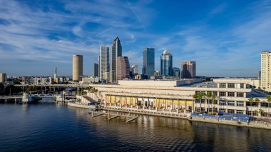 Tampa, Florida 'nın hava manzarası modern yüksek binalarla dolu canlı bir ufuk çizgisi, şehir merkezinden geçen ikonik Hillsborough Nehri, Tampa Körfezi boyunca geniş deniz kenarları ve yemyeşil noktalı kentsel alanlar gösteriyor..
