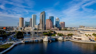 Tampa, Florida 'nın hava manzarası modern yüksek binalarla dolu canlı bir ufuk çizgisi, şehir merkezinden geçen ikonik Hillsborough Nehri, Tampa Körfezi boyunca geniş deniz kenarları ve yemyeşil noktalı kentsel alanlar gösteriyor..