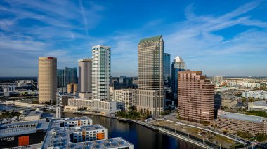 Tampa, Florida 'nın hava manzarası modern yüksek binalarla dolu canlı bir ufuk çizgisi, şehir merkezinden geçen ikonik Hillsborough Nehri, Tampa Körfezi boyunca geniş deniz kenarları ve yemyeşil noktalı kentsel alanlar gösteriyor..