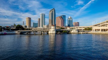 Tampa, Florida 'nın hava manzarası modern yüksek binalarla dolu canlı bir ufuk çizgisi, şehir merkezinden geçen ikonik Hillsborough Nehri, Tampa Körfezi boyunca geniş deniz kenarları ve yemyeşil noktalı kentsel alanlar gösteriyor..
