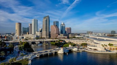 Tampa, Florida 'nın hava manzarası modern yüksek binalarla dolu canlı bir ufuk çizgisi, şehir merkezinden geçen ikonik Hillsborough Nehri, Tampa Körfezi boyunca geniş deniz kenarları ve yemyeşil noktalı kentsel alanlar gösteriyor..