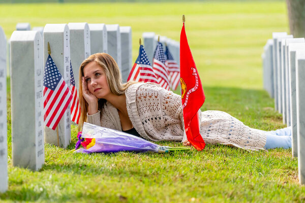 A young bride kneels at a military cemetery amid uniform rows of graves. Clutching flowers and a tombstone, tears streak her face as she mourns a beloved family member with profound, silent grief.