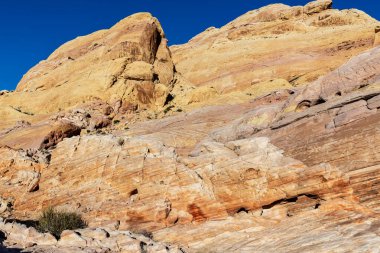Nevada çölünden yükselen görkemli kaya oluşumları, güneşin altında parlayan engebeli dokuları, kurak arazinin ham güzelliğini vurgulayan engin mavi gökyüzüne karşı..