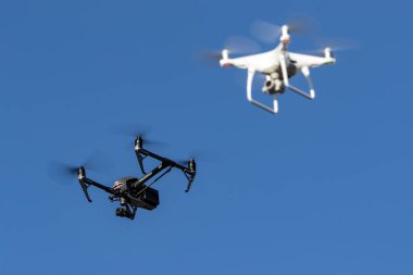 Her boyutta ve ağırlıkta çeşitli bir dron filosu açık mavi gökyüzünde süzülür. Parlak çerçeveleri, senkronize uçuşlarda zarifçe manevra yaparken güneş ışığında parlar..