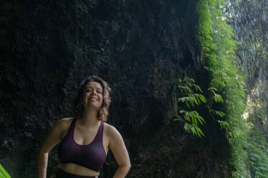 Spor kıyafetli genç bir kadın doğa manzarasında duruyor, kameraya bakıyor ve gülümsüyor, Bali