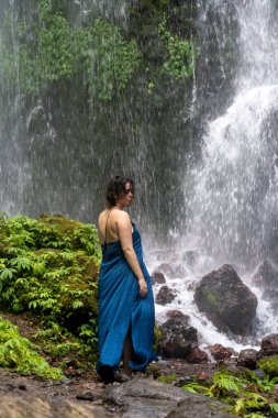 Mavi bir elbiseyle şelalenin önünde duran güzel bir kadının portresi. Bali. Endonezya.