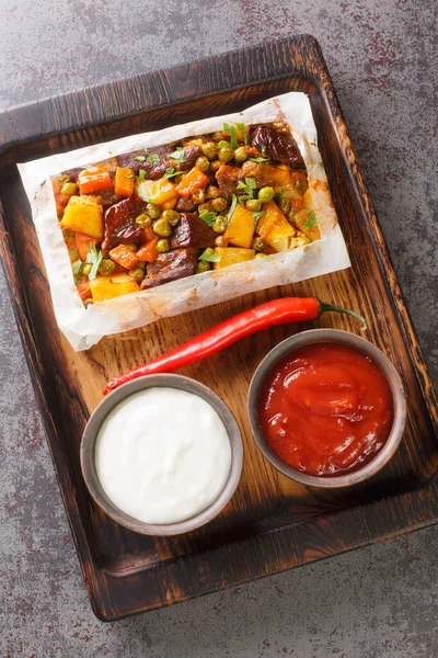 Turkish kebab with vegetables in parchment, accompanied by sauces close-up on a wooden tray on the table. Vertical top view from abov