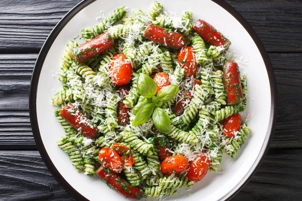 Hearty lunch Fusilli pasta with sausages, tomatoes, green pesto sauce and cheese close-up in a plate on the table. Horizontal top view from abov