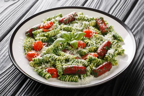 Hearty lunch Fusilli pasta with sausages, tomatoes, green pesto sauce and cheese close-up in a plate on the table. Horizonta