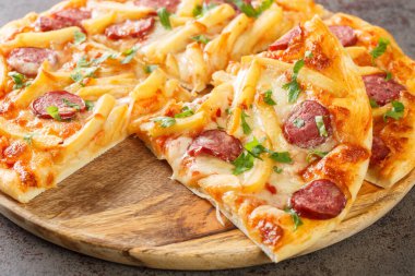 Sliced pizza with french fries, sausages, mozzarella cheese, tomato sauce and herbs close-up on a wooden board on the table. Horizonta