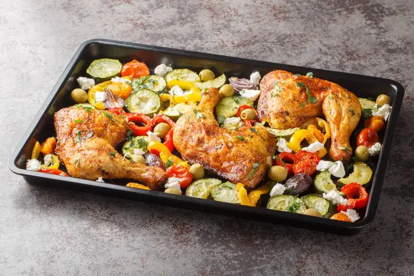Greek style baked chicken quarters with zucchini, tomatoes, peppers, olives and feta cheese close-up on a baking sheet on the table. Horizonta