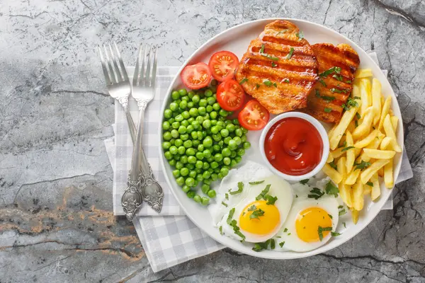 Fried pork loin steak served with French fries, fied eggs, green pea and fresh tomato closeup on the plate on the table. Horizontal top view from abov