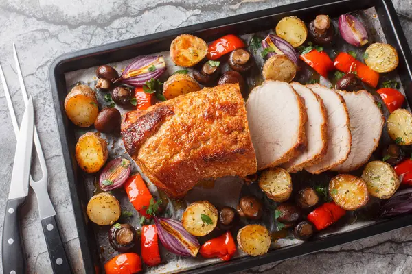 Honey baked pork loin with potatoes, onions, bell peppers and mushrooms close-up on a baking sheet on the table. Horizontal top view from abov