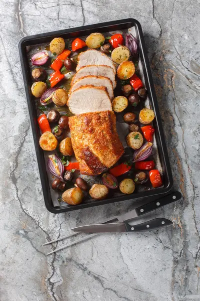 Baked pork loin with vegetables and mushrooms close-up on a baking sheet on the table. Vertical top view from abov