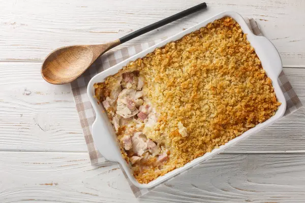 Hot Chicken Cordon Bleu Casserole has layers of chicken, ham, cheese sauce, and buttery Panko breadcrumbs close up in the baking dish on the white wooden table. Horizontal top view from abov