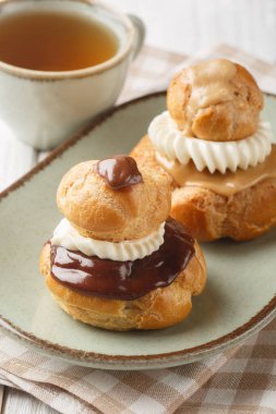 Yumuşak vanilyalı pasta kremiyle doldurulmuş ve koyu çikolata ganacıyla kaplanmış dini choux hamur tatlısı çörekleri ya da ahşap masadaki tabağa yakın kahve. Dikey