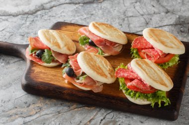 Delicious Italian Tigelle sandwich with salami, ham, olive, tomato, lettuce and arugula close-up on wooden board on table. Horizonta clipart