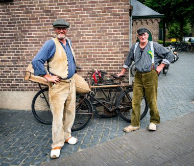 Lunteren, 25-aug-2023: Geleneksel Hollanda giysileri içinde eski lunterse dagen üzerinde eski moda bisikletlerle tahta takunyalı iki adam