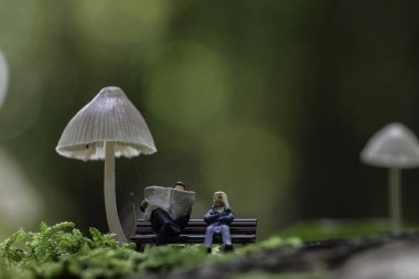 tiny figures from little world sit on a bench beneath oversized mushrooms in a creative concept scene. The image evokes a whimsical and imaginative ambiance, clipart