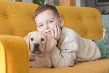 A boy in a knitted sweater poses on a yellow sofa with his Labrador puppy