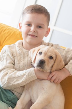 A boy in a knitted sweater poses on a yellow sofa with his Labrador puppy