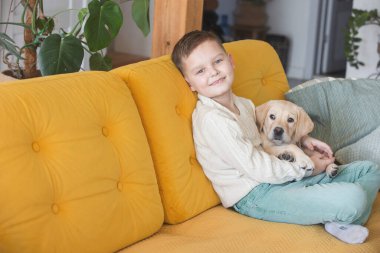 A boy in a knitted sweater poses on a yellow sofa with his Labrador puppy