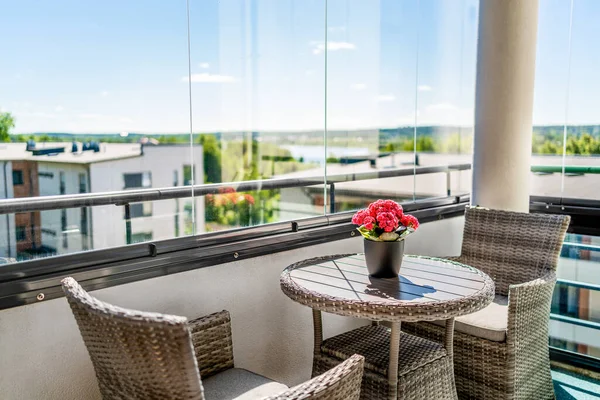 Balcony in summer. Home patio with a view. Condo apartment in city. Scandinavian decor and furniture design. Glass windows, glazing. Flower or plant on table and chairs. Sunny terrace in modern flat.