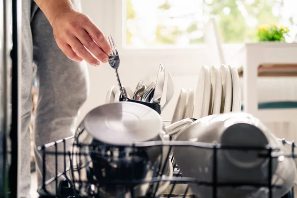 Dish washer machine in kitchen. Man loading dishwasher. Washing plates. Fork in hand. Full of cutlery. Clean or dirty. Pot and tableware. Household chores and housework in family. Unloading utensils.