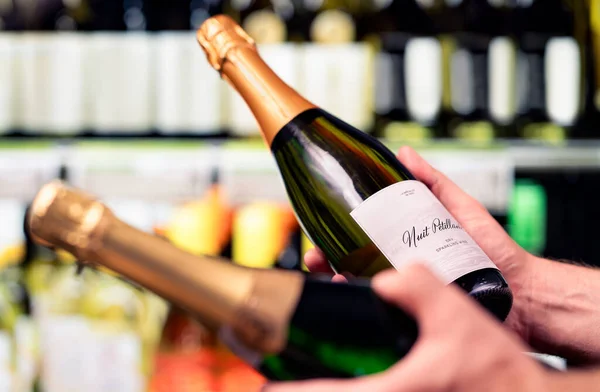 Sparkling wine in alcohol store. Champagne or prosecco in liquor shop. Man holding two bottles in hands, choosing drink. Customer compare products in spirits aisle in supermarket. Mockup brand label.