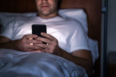 Geceleri yatakta telefon. Uyumadan önce akıllı telefonu olan bir adam. Gizli mesajlaşma, hile yapma ya da geç saatlere kadar çalışma. Mutlu adam internette haberlere bakıyor. Cep telefonunu elinde tutuyorsun. Karanlık ev yatak odası. Ekran zaman kavramı.