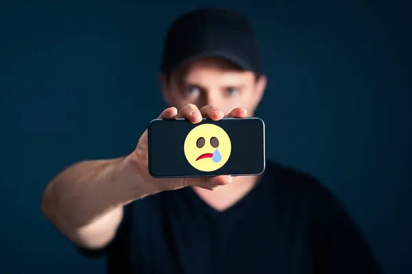 Sad unhappy emotion icon in phone. Depressed lonely man with smartphone. Online hate, trauma, cyber bullying or social media pressure concept. Male mental health taboo. Internet harassment victim.