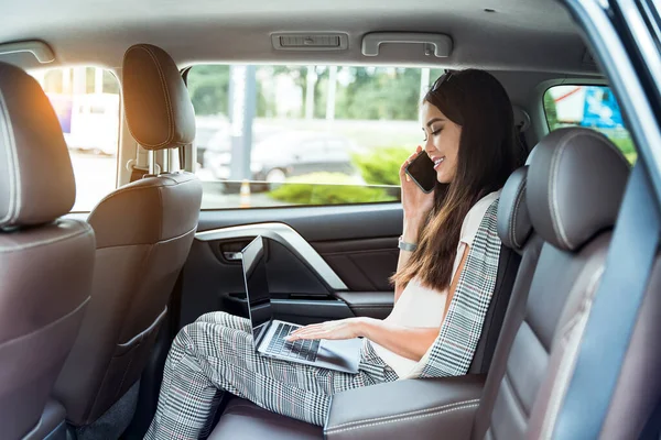 Young woman boss, top manager sitting in the back seat of luxury car and talking phone while working on laptop.