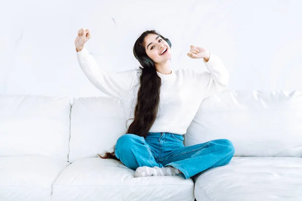 Pretty indian girl in wireless headphones listening to music and dancing while sitting on white leather couch in white interior