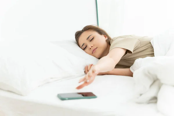 Morning awakening with alarm clock in the phone. Sleepy girl reaches for her smartphone on the bed.