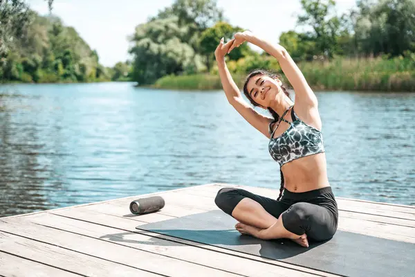 Young Indian woman training yoga outdoors sitting on pier by the river.