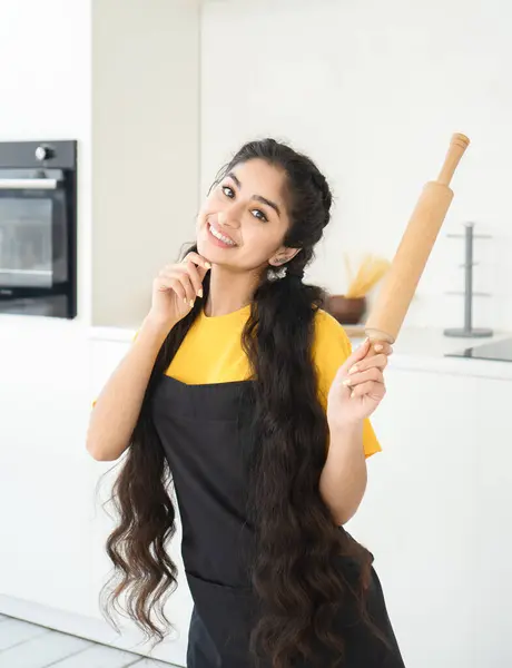 Beautiful Indian woman in kitchen robe and pastry rolling pin in hand poses in modern kitchen