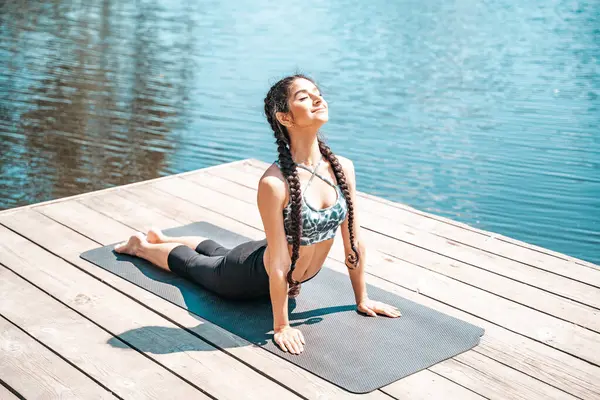 Young indian woman doing cobra pose from yoga lying on pier by the river.