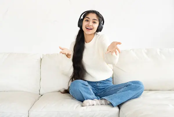 Cheerful Indian girl in wireless headphones sitting on leather couch
