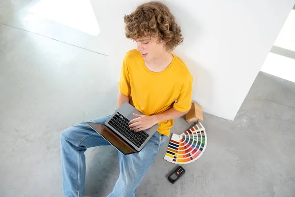 Interior designer works on project on laptop sitting on the floor. Next to him lies a fan of sample colors and a laser tape measure. Interior of the room is prepared for finishing with finishing materials.