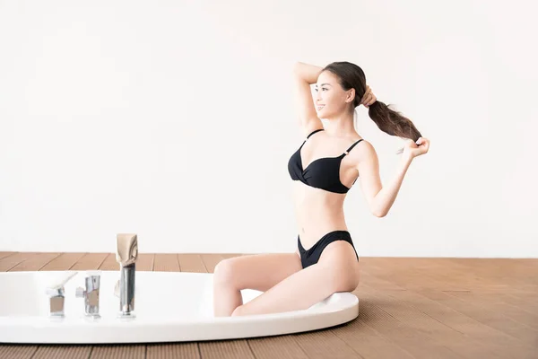Beautiful Korean girl in black swimsuit sitting on round bubble bath in modern interior.