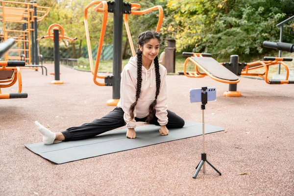 Online fitness training in modern outdoors sportground performed by young Indian woman doing twine and looking at smartphone