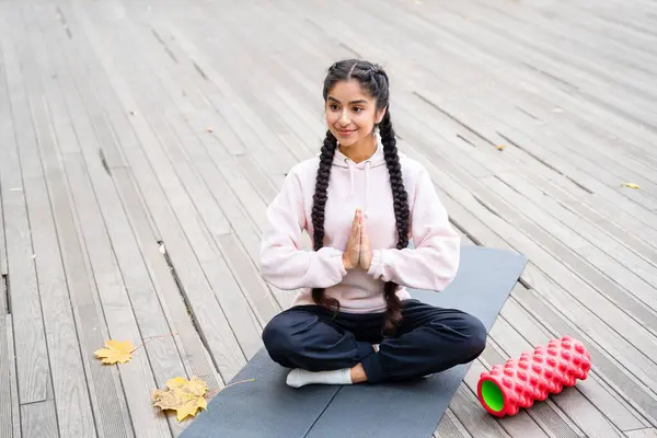 Young Indian woman practicing yoga pilates in the autumn park, sitting on fitness mat