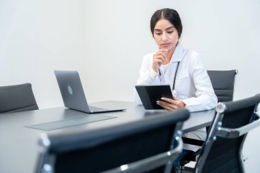 Modern ofiste beyaz laboratuvar önlüğü giyip dijital tablette hastanın tıbbi geçmişini okuyarak odaklanmış bir ifadeyle yorgun düşmüş bir doktor..