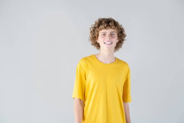 Parlak sarı tişörtle poz veren kıvırcık saçlı ve sıcak gülüşlü yakışıklı bir genç. Çekicilik ve güven saçarak doğrudan kameraya bakıyor, dostane ve davetkar bir portre oluşturuyor..