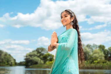 Geleneksel turkuaz giyinmiş güzel Hintli kadın doğal arka plana karşı zarif bir şekilde Namaste jesti ile poz veriyor..