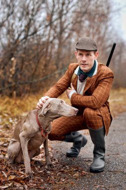 caucasian male with pet dog going to hunt together in wild nature in countryside, young american guy in elegant brown suit and hat with rifle shotgun. hunting, animals, wildlife concept