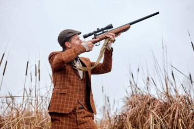 confident hunter wearing brown fashionable suit and hat waiting for the bird, aiming rifle shotgun up at sky. concentrated caucasian Hunter on hunt. Illegal Hunting Birds. Hunting without borders