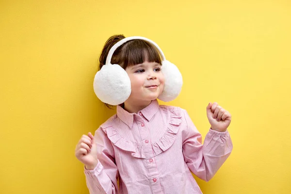 Cute caucasian child girl in blouse listen to music in headphones and dance, wearing big wireless earphones isolated on yellow studio background