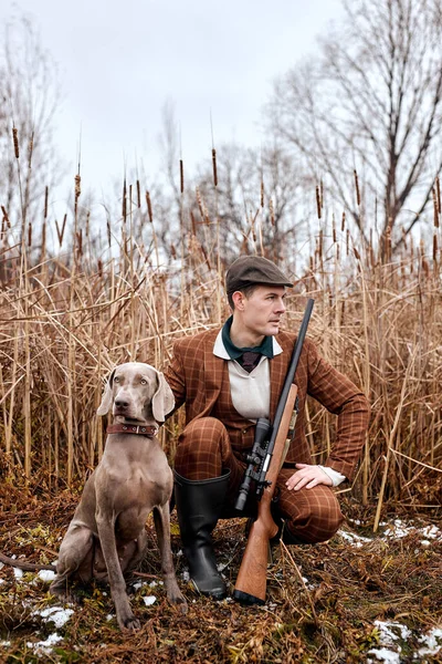 Serious caucasian hunter man with dog holding shotgun rifle walking in countryside nature, wearing brown trendy stylish suit and hat. young skilled experienced male hunter waiting for prey